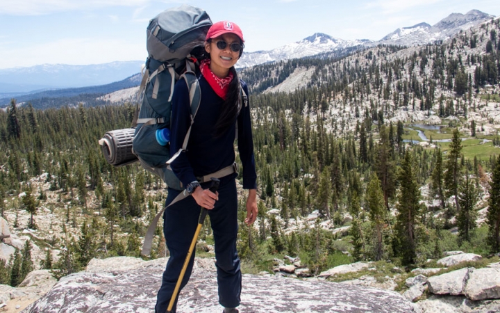 a person carrying a backpack smiles at the camera with a mountain landscape in the background 